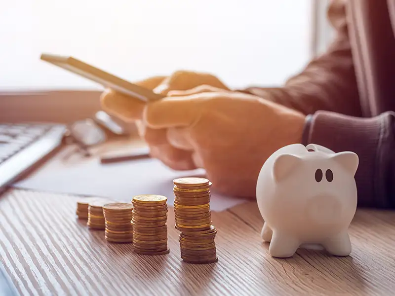 Person using their phone on a desk with coins and a piggybank
